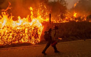 Firefighter walking while holding tools with a red blazing wildfire in the background