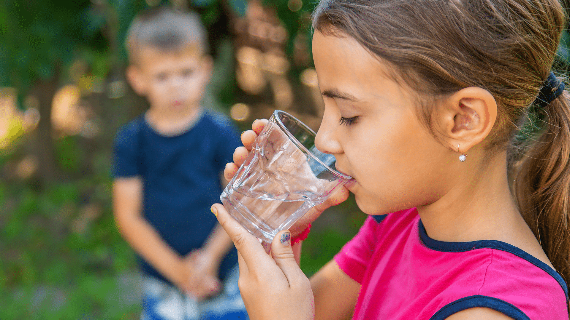 A child drinks water from a clear plastic cup with another child in the background